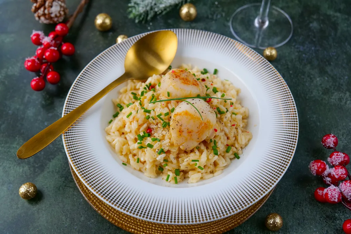 Assiette blanche avec un risotto crémeux, trois noix de Saint-Jacques dorées au centre et zeste de citron, posé sur une table en bois clair